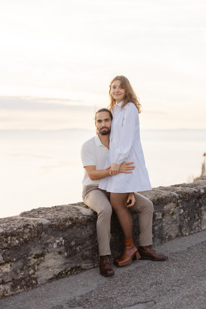 Bride and groom sitting on stone wall with Lake Geneva behind during engagement session Lavaux