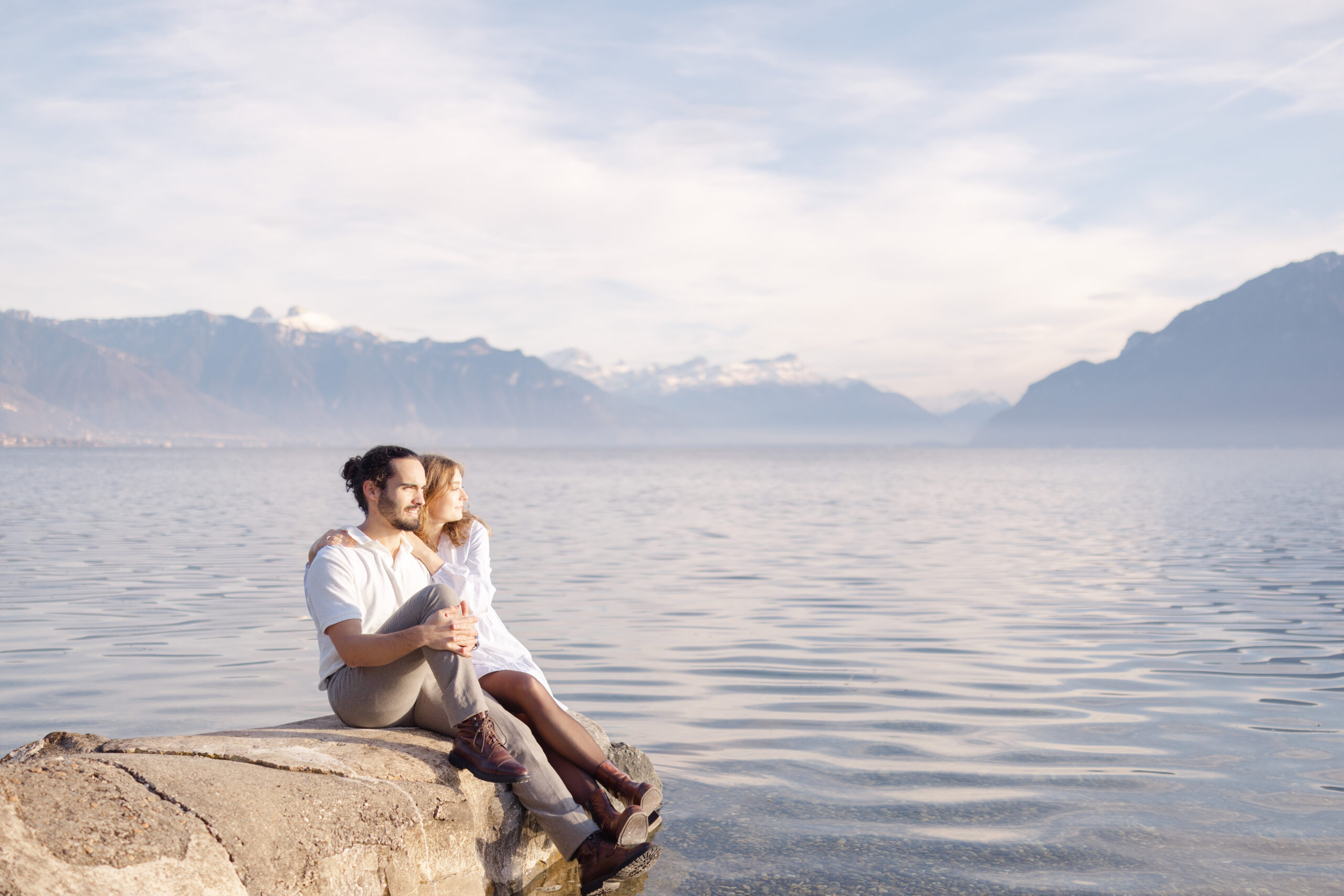 Couple sitting on rock by Lake Geneva with Swiss Alps in background, engagement photo session Lavaux